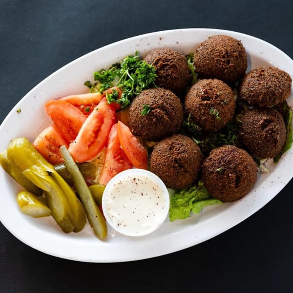 A white oval plate of falafel, sliced tomatoes, pickles, and a side of white sauce.