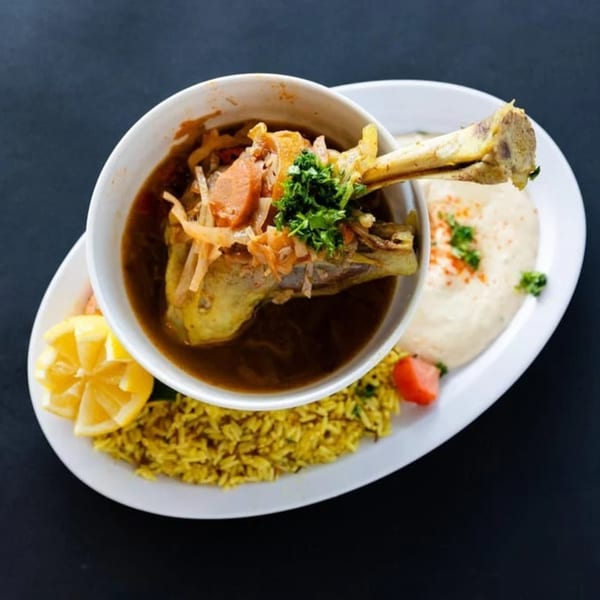 A Sultan's Restaurant lamb shank stew with rice, hummus, and lemon wedges on a white plate.