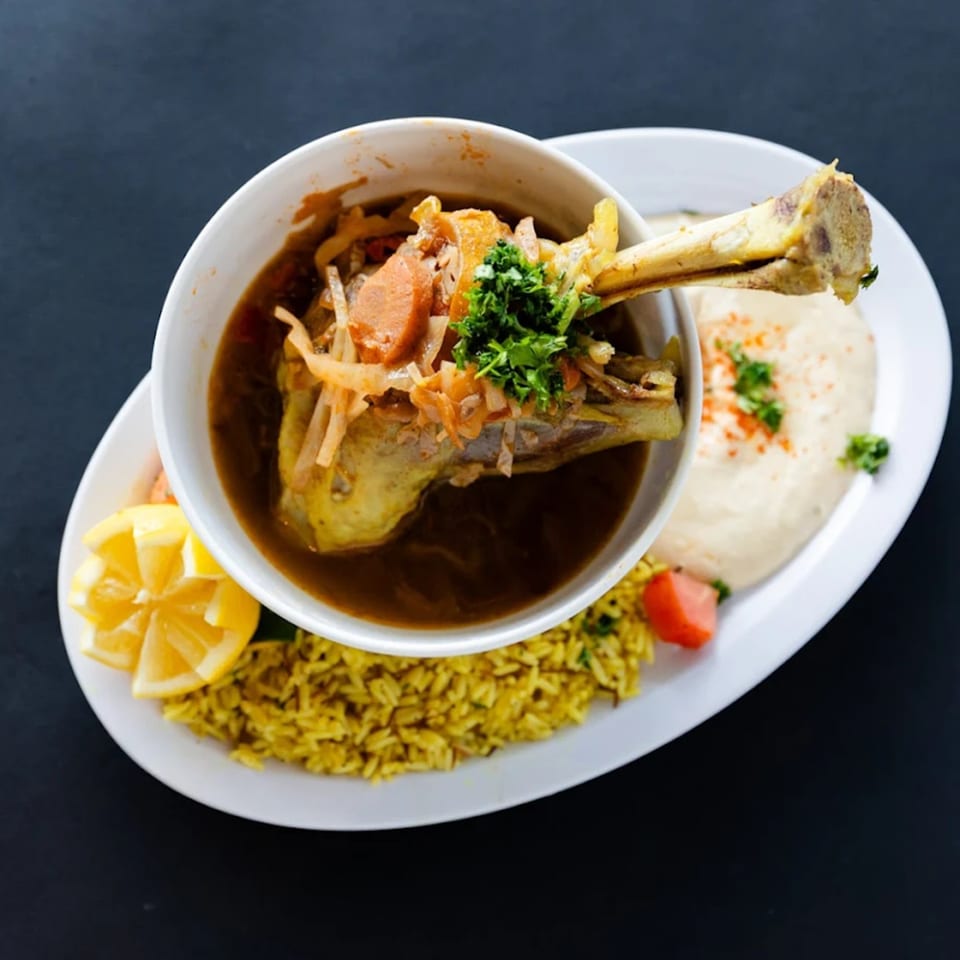 A Sultan's Restaurant lamb shank stew with rice, hummus, and lemon wedges on a white plate.