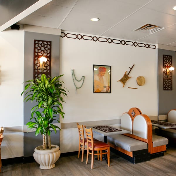 Mediterranean restaurant interior with booths, wooden chairs, potted plant, and wall decor including a framed painting.