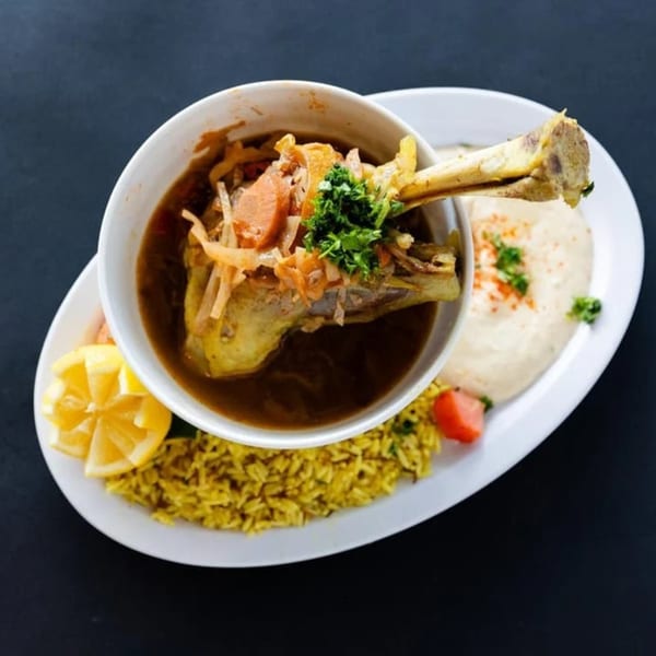 Mediterranean lamb shank stew with rice, hummus, and lemon wedges on a white plate.
