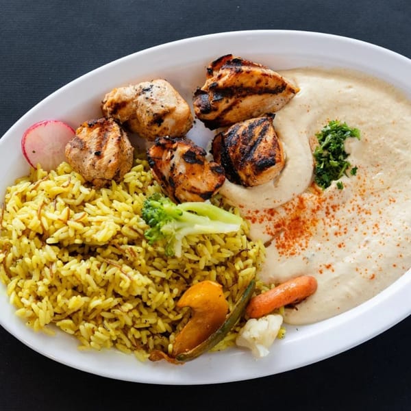 Grilled chicken skewers, yellow rice, hummus, and vegetables on a white oval plate.